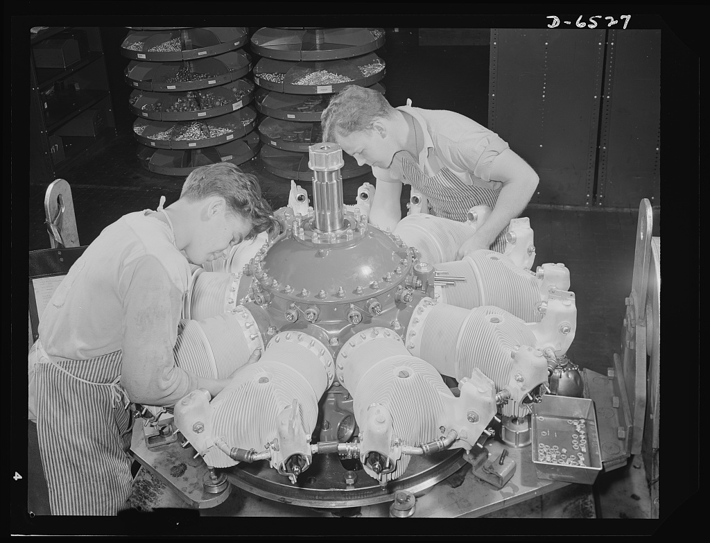Two young men with striped aprons are bent over an airplane engine in a factory. The photo is in black and white