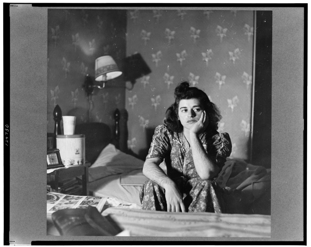 A young woman sits on a bed with her head in one hand. She is in a small room with a cluttered nightstand, two twin beds, patterned wallpaper, and a small lamp. The photo is in black and white.