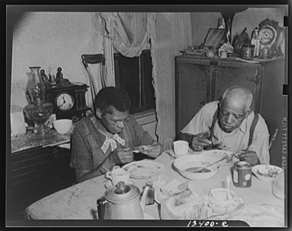 An old couple sit at a cluttered kitchen table eating together. Behind them in the kitchen is a low counter with a clock and vase and a taller cabinet with another clock and various trinkets. The photo is in black and white.
